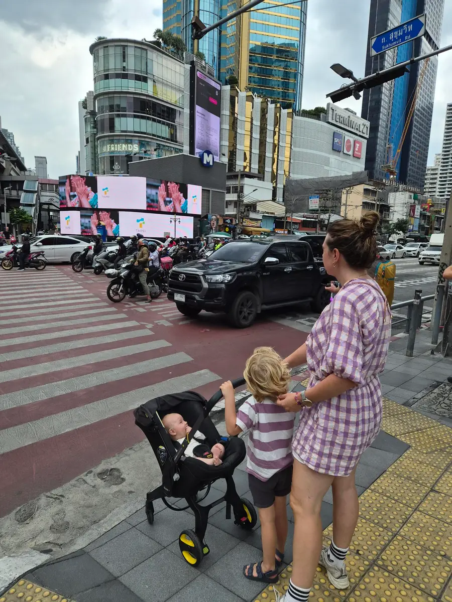 A mother and an older child wait to cross Sukhumvit Road in Bangkok, with a baby in a Doona car seat stroller; the Terminal 21 mall is behind them.